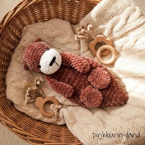 May include: A handmade, plush, brown teddy bear with a white face and black details, lying on a cream-colored knitted blanket. Two wooden baby teethers and dried flowers are also in the wicker basket. The text "pinkbunnyland" is visible.