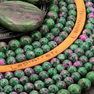 Peut inclure: Gros plan sur un collier de perles de pierres précieuses vertes et roses. Les perles sont rondes et ont un aspect moucheté. Les perles sont enfilées sur un cordon de cuir brun avec le mot "Lapierre" en relief.