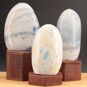 Peut inclure: Trois pierres d'agate bleu et blanc polies, chacune avec un motif unique, sont présentées sur des socles en bois. Les pierres sont lisses et ont une forme naturelle et organique.