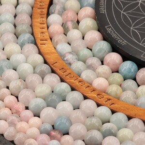 May include: A close-up of a strand of light blue, pink, and white gemstone beads. The beads are round and smooth, and they are arranged in a circular pattern. The beads are on a wooden surface with the word "Lapierreft" written on it.