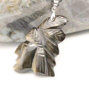 Peut inclure: Un pendentif en obsidienne noire sculpté en forme de chef amérindien portant une coiffe. Le pendentif est détaillé avec des sculptures complexes et a une bélière en argent.