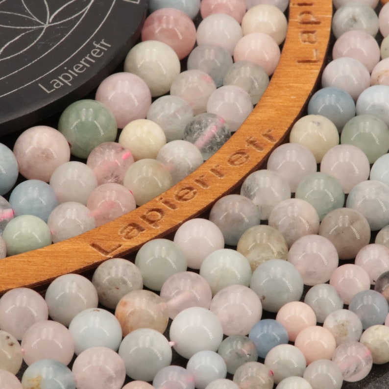 May include: A close-up of a strand of pastel-colored gemstone beads. The beads are round and smooth, and they are arranged in a circular pattern. The beads are a mix of light blue, pink, and white.