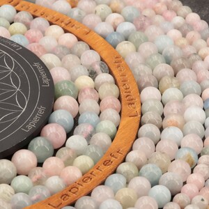 May include: A close-up of a large quantity of pastel-colored gemstone beads. The beads are arranged in a circular pattern around a wooden ring with the word "Lapierreff" inscribed on it.