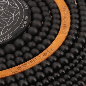 Peut inclure: Gros plan sur un cercle en bois avec le mot "Lapierreff" gravé dessus. Le cercle est entouré de plusieurs rangées de perles noires.