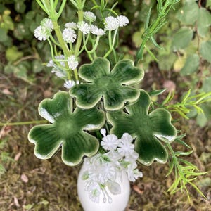 Pode incluir: Um vaso de cerâmica branca contendo três ornamentos de trevo de quatro folhas verde-escuro brilhante em hastes, intercalados com ramos de pequenas flores artificiais brancas e folhagem verde fina, criando um arranjo botânico decorativo.