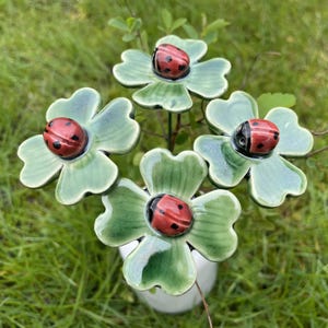 May include: A decorative garden stake featuring four ceramic four-leaf clovers in shades of light and dark green. Each clover is topped with a small, glossy red ladybug figurine with black spots. The clovers are arranged in a cluster on a stem.