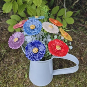 May include: A white ceramic pitcher containing a bouquet of artificial flowers with multi-colored ceramic petals in shades of blue, red, purple, orange, and cream, each featuring a textured yellow center, accented by small white filler flowers.