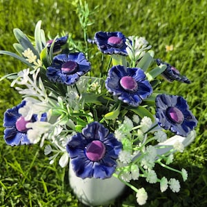 Puede incluir: Un ramo de flores de cerámica en un jarrón blanco. Las flores son de un azul profundo con centros morados. El follaje blanco y verde complementa el arreglo, sobre un fondo de hierba verde.