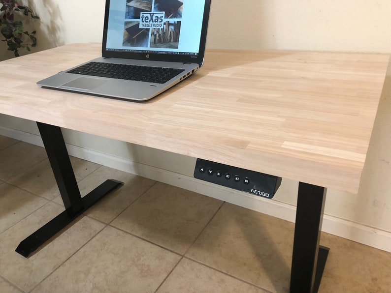 May include: A light wood standing desk with a black frame and a black control panel labeled "FEITUO". A laptop computer is on the desk.