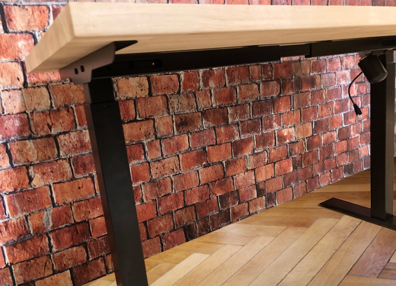 May include: Black adjustable standing desk frame with a light wood desktop. The frame has a black metal base and a black metal support beam. The desk is in front of a brick wall.