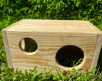 Könnte beinhalten: Eine rechteckige, helle Holzkiste mit zwei unterschiedlich großen, kreisförmigen Öffnungen auf der Vorderseite. Die Holzmaserung ist auf der gesamten Oberfläche der Kiste sichtbar, die eine flache Oberseite und massive Seitenwände aus Sperrholz aufweist.