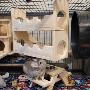 May include: A gray chinchilla sits on a wooden platform with a small wooden picnic table and a food bowl. The chinchilla is in a cage with a wooden play structure and a black metal wheel.