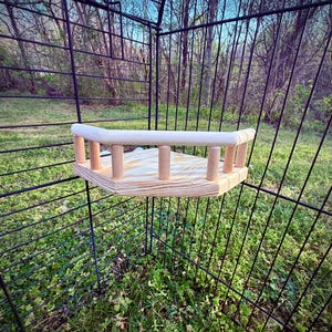 May include: A wooden corner perch for a pet cage. The perch has a light-colored, unfinished wood platform with a curved top rail and vertical dowels for support. The perch is set inside a black wire cage, with a background of green grass and trees.