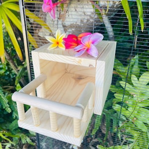 May include: A wooden birdhouse with a small balcony and a removable front panel. The birdhouse is attached to a black wire cage and is surrounded by pink and yellow flowers.