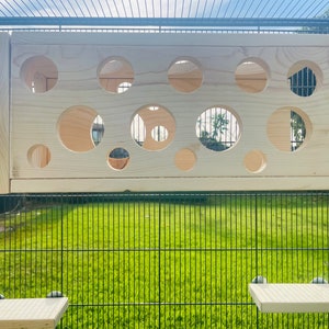 May include: A wooden play structure for small animals, featuring a panel with multiple circular openings of varying sizes. The panel is mounted inside a black wire cage.