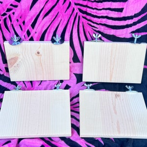 May include: Four unfinished wooden shelves with metal brackets. The shelves are all the same size and are arranged in a row. The shelves are on a pink and black patterned background.