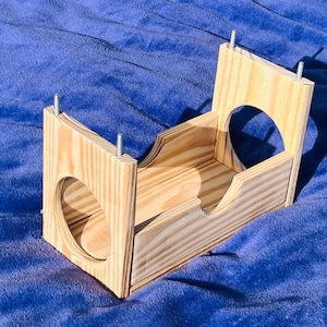 May include: A wooden pet bed with a tunnel entrance and a raised platform. The bed is made of natural wood and has a simple design.