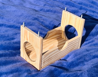 Puede incluir: Una cama de madera para mascotas con una entrada de túnel y una plataforma elevada. La cama está hecha de madera natural y tiene un diseño simple.