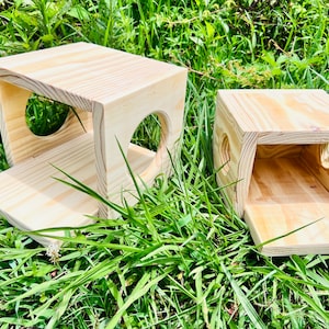 May include: Two wooden boxes with openings on the sides, designed for small animals. The boxes are made of natural wood and have a light brown color. They are sitting on a bed of green grass.