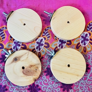 Puede incluir: Cuatro juguetes masticables de madera para mascotas con herrajes metálicos. Los juguetes son redondos y tienen un agujero en el centro. Están sobre una tela estampada de color rosa y morado.