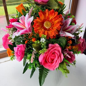 May include: A colourful floral display with pink roses, orange gerberas, and pink lilies. The arrangement includes various green foliage and small white flowers, creating a full and vibrant display. The flowers are arranged in a compact, eye-catching design.