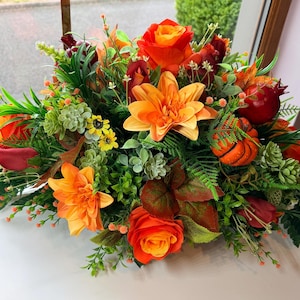 May include: A floral arrangement featuring orange roses, orange lilies, and orange dahlias mixed with small yellow flowers, green succulents, fern fronds, and red pomegranate-like accents. The composition includes various green leaves and foliage.