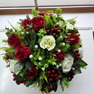 May include: A circular floral display with red and white roses, and red berries. The arrangement is complemented by green foliage and small white blossoms. The flowers are densely packed, giving a full, abundant look.
