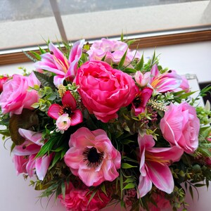 May include: A colourful floral display with pink and fuchsia blooms, such as roses, lilies, and peonies, and green leaves. The arrangement is set against a bright background, highlighting the different textures and colours.