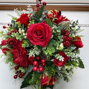 May include: A lush floral display with deep red roses, dahlias, and berries. The arrangement is complemented by green foliage and small white flowers, creating a full, textured look. A small pine cone adds a natural element to the design.