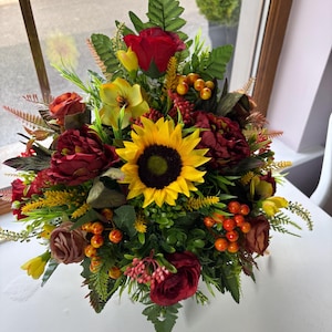 May include: A colourful floral display with a prominent sunflower, deep red roses, and yellow and burgundy blooms. Green leaves and orange berries provide contrast. The arrangement is presented on a white surface.