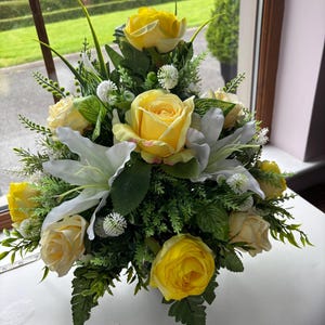 May include: A vibrant floral display with yellow roses and white lilies, complemented by green leaves and small white blooms. The arrangement is positioned near a window, offering a view of a green, manicured landscape.
