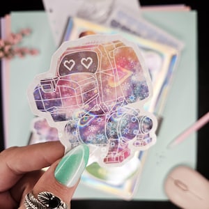 May include: A clear sticker with a holographic finish featuring a cartoon robot with a galaxy design. The robot has two hearts for eyes and is holding a weapon. The sticker is on a white background.