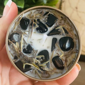 Op de afbeelding: Een witte kaars met zwarte stenen en gedroogde kruiden ingebed in de was. De kaars zit in een gouden blik.