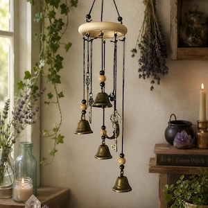 Op de afbeelding: Een hangende windgong met messing bellen, houten kralen en decoratieve sleutels. De windgong is opgehangen aan een lichtgekleurde houten ring en zwarte koorden. De bellen zijn verschillend van grootte en hangen op verschillende lengtes.