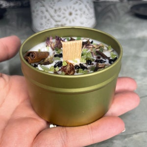 May include: A gold metal tin candle with a white wax top. The candle is filled with dried herbs, green and black crystals, and a wooden wick.