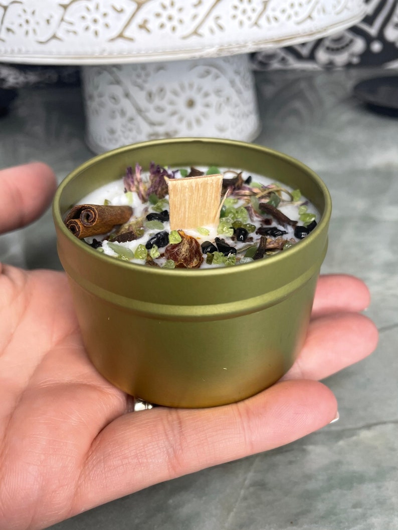 May include: A gold metal tin candle with a white wax top. The candle is filled with dried herbs, green and black crystals, and a wooden wick.