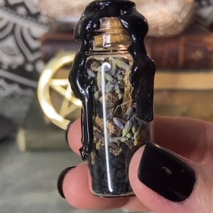 May include: A small glass bottle filled with dried herbs and black crystals, sealed with black wax. The bottle is held in a hand with black nail polish.