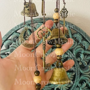 Op de afbeelding: Een hangende windgong met bronskleurige bellen, sleutels en een maansikkelsieraad. De windgong bevat houten kralen en is opgehangen aan bruine koorden. Het ontwerp heeft een vintage esthetiek.