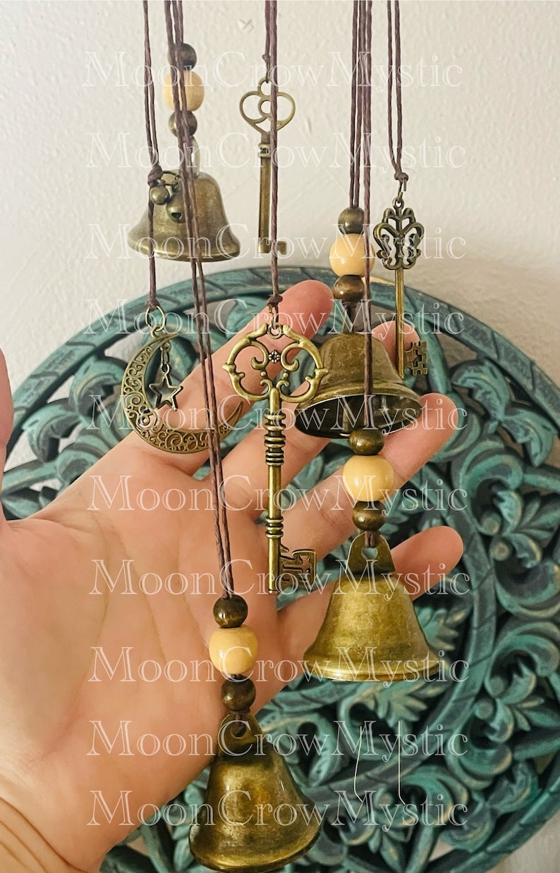 Op de afbeelding: Een hangende windgong met bronskleurige bellen, sleutels en een maansikkelsieraad. De windgong bevat houten kralen en is opgehangen aan bruine koorden. Het ontwerp heeft een vintage esthetiek.
