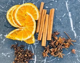 Op de afbeelding: Gedroogde sinaasappelschijfjes, kaneelstokjes, steranijs en kruidnagels op een grijs marmeren oppervlak. Deze ingrediënten worden vaak gebruikt in kerstbakkerij en dranken.