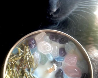 Könnte beinhalten: Eine Kerze in einer goldfarbenen Dose, gefüllt mit weißem Wachs, bunten Steinen und Rosmarinzweigen. Die Kerze befindet sich vor einer schwarzen Katze mit grünen Augen.