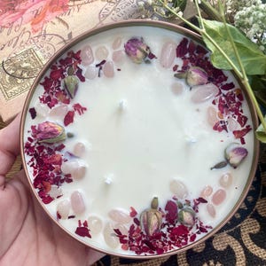 May include: A white candle with a copper-colored container. The candle is decorated with rose petals and pink crystals around the edge.