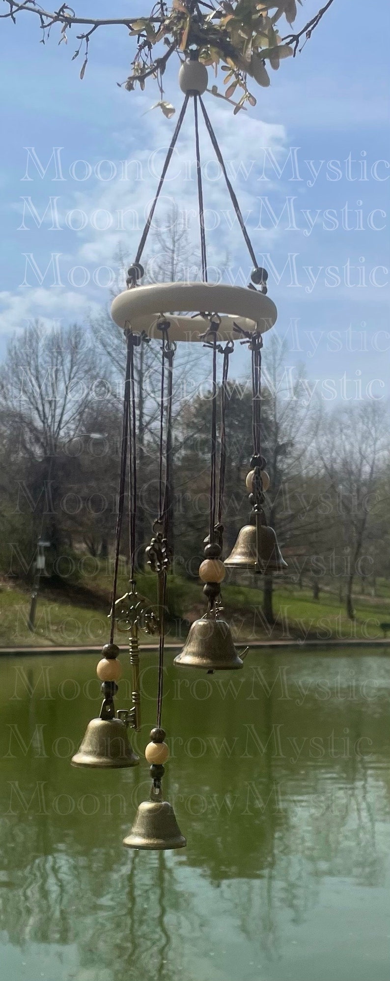 Op de afbeelding: Een hangende windgong met een cr&egrave;mekleurige houten ring en kleine bronzen bellen. De windgong bevat decoratieve sleutels en houten kralen, opgehangen aan een tak tegen een achtergrond van lucht en water.