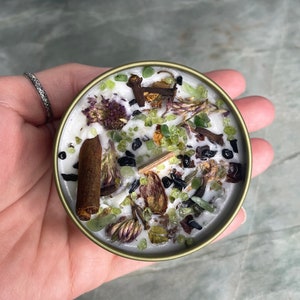 May include: A white candle with green and purple dried flowers, black and brown spices, and green and black crystals embedded in the wax. The candle is in a gold metal container.