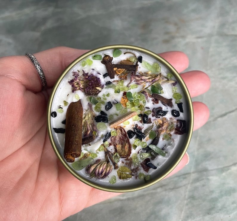 May include: A white candle with green and purple dried flowers, black and brown spices, and green and black crystals embedded in the wax. The candle is in a gold metal container.