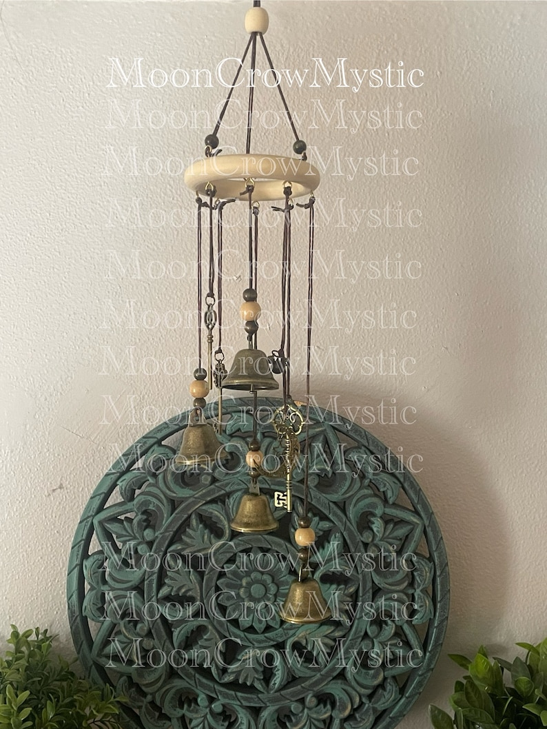 Op de afbeelding: Een hangende windgong met messing bellen, houten kralen en kleine sleutels. De windgong is opgehangen aan een cr&egrave;mekleurige houten ring. De achtergrond toont een turquoise, gebeeldhouwde houten wanddecoratie.