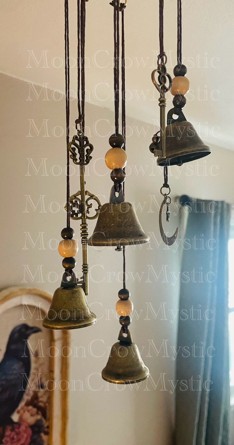 Op de afbeelding: Een hangende windgong met bronzen bellen, decoratieve sleutels en houten kralen. De bellen hangen aan bruine koorden, met een maansikkelsieraad aan &eacute;&eacute;n ervan. De achtergrond is een zachte, neutrale kleur.