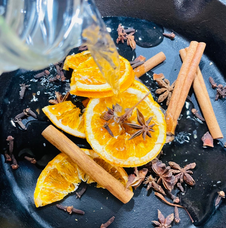 Puede incluir: Una olla de hierro fundido negra llena de agua, rodajas de naranja, palitos de canela, an&iacute;s estrellado y clavos. El agua se vierte desde una jarra transparente.
