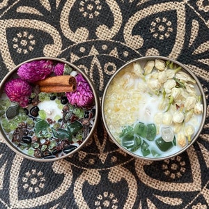 May include: Two candles with different scents and ingredients. The first candle has a brown, black, and green color scheme with dried flowers, cinnamon sticks, and green stones. The second candle has a white and green color scheme with white flowers and green stones.