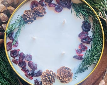May include: A white candle with a gold rim, decorated with dried cranberries, pine sprigs, and pine cones. The candle is in a round glass jar.